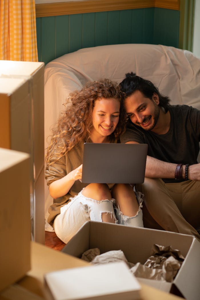 Smiling couple using laptop while unpacking boxes in their new home. Bright, cozy indoor setting perfect for relocation themes.